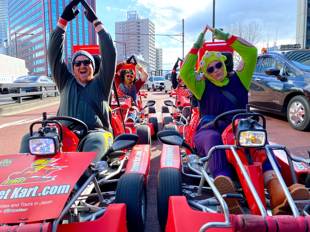 Group of friends in red go-karts on a city street, two in foreground making triangle shapes with arms; one wears a frog hood costume and sunglasses.