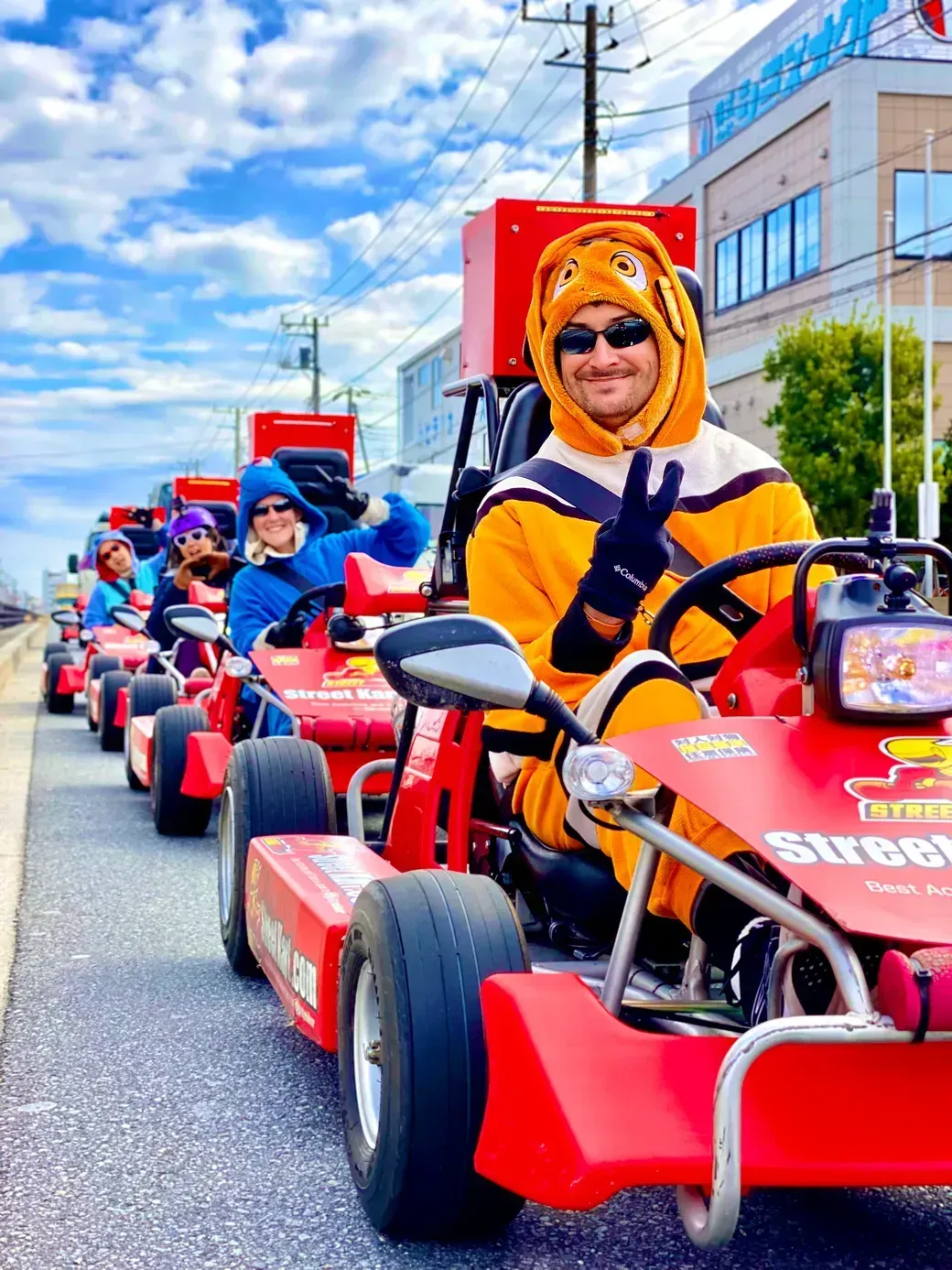 Man in an orange mascot suit driving a red go-kart in a street parade, flashing a peace sign; riders in colorful outfits follow behind.
