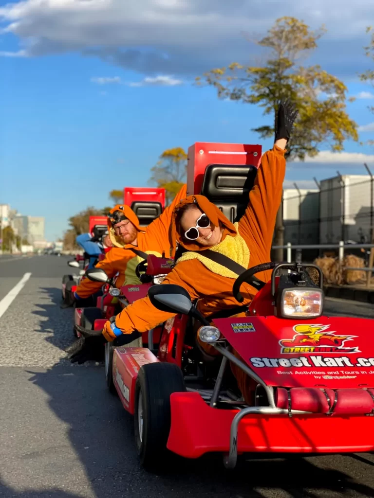 People in orange jumpsuits riding red go-karts in a parade, one driver leaning out with a raised arm and white sunglasses.